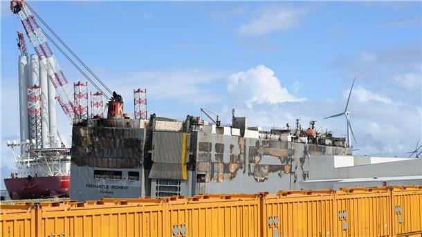 Der schwer beschädigte Autofrachter „Fremantle Highway“ liegt im Hafen, abgeschirmt durch gelbe Container.