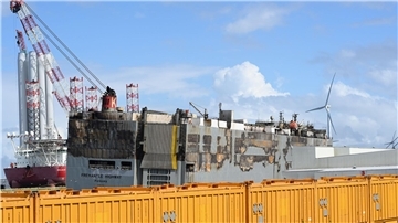 Der schwer beschädigte Autofrachter „Fremantle Highway“ liegt im Hafen, abgeschirmt durch gelbe Container.