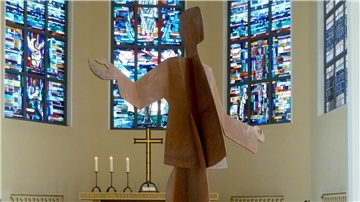 ALT-Text:
Skulptur „Tanzender Christus“ aus Stahl in der Großen Kirche Bremerhaven vor farbigen Kirchenfenstern mit biblischen Motiven und einem Altar mit drei brennenden Kerzen.