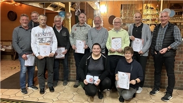 Eine Gruppe von elf Personen, überwiegend Männer, posiert in einem gemütlich dekorierten Raum mit Luftschlangen und Ballons. Mehrere Personen halten Urkunden in den Händen.