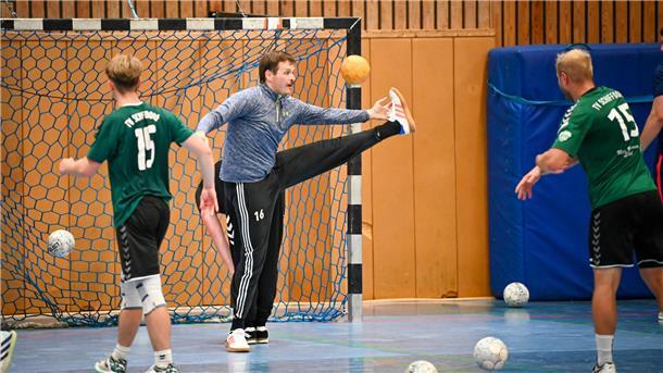 Ein Handball-Torwart steht während des Trainings im Tor und wehrt die Bälle zweier Mitspieler ab. Hinter dem Torhüter bückt sich ein weiterer Spieler.