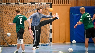 Ein Handball-Torwart steht während des Trainings im Tor und wehrt die Bälle zweier Mitspieler ab. Hinter dem Torhüter bückt sich ein weiterer Spieler.