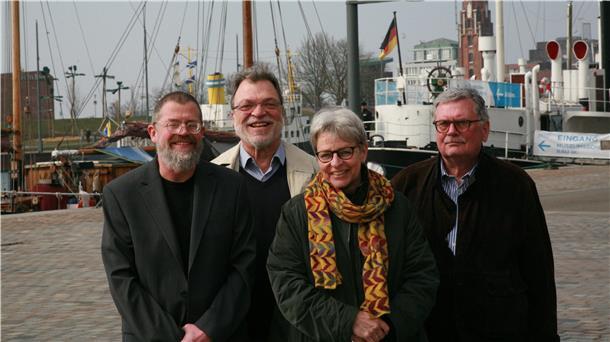 Drei Männer und eine Frau, alle zwischen Ende 50 und Anfang 70, blicken in die Kamera. Sioe stehen vor einem Hafenbecken, im Hintergrund sind alte Segelschiffe zu erkennen.