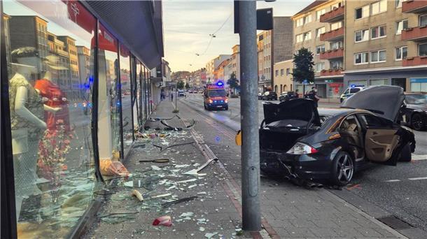 Glasscherben von einem Schaufenster liegen auf einem Fußweg. Daneben steht ein stark beschädigter schwarzer Wagen.