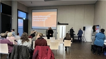 Ein Konferenzraum, in dem mehrere Menschen sitzen und auf einen Bildschirm schauen, auf dem „Herzlich willkommen zur Innovationswerkstatt Diversity in KMU“ steht.