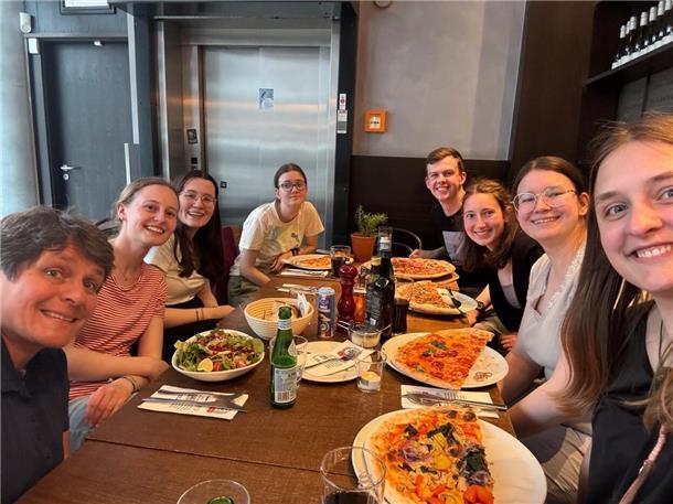 Frauen und Männer sitzen mit Pizza und Salat auf Tellern an einem Tisch und lachen in die Kamera.