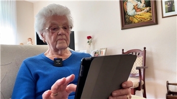 Die 85-jährige Hilde Heuer sitzt in einem blauen Pullover auf einem Sofa und liest etwas auf ihrem Tablet.
