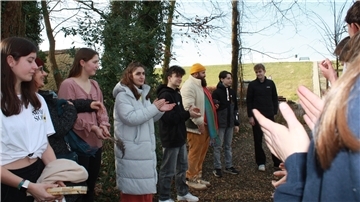 Eine Gruppe junger Menschen steht im Halbkreis im Freien, umgeben von Bäumen und Natur. Einige klatschen, während andere aufmerksam zuhören.