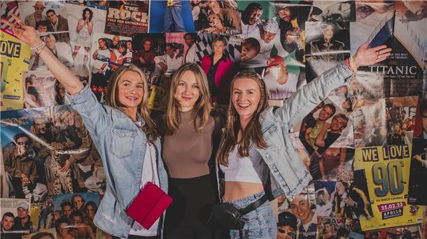 Drei junge Frauen vor einer Wand mit Plakaten der 90er-Jahre.