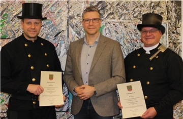 Drei Männer posieren vor einer bunt gemusterten Wand. Zwei von ihnen tragen die traditionelle schwarze Schornsteinfeger-Uniform mit Zylinder und halten ihre Bestellungsurkunden in der Hand. Der Mann in der Mitte trägt ein graues Sakko und gratuliert den beiden.