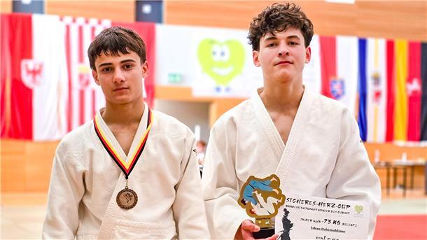 Zwei junge Judoka blicken in die Kamera, ein Athlet trägt eine Bronzemedaille um den Hals, der andere hält eine Urkunde und einen Sierpokal.