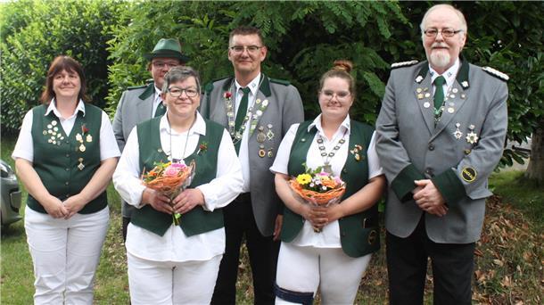 **ALT-Text:**
Sechs Mitglieder des Schützenvereins Gackau stehen in Uniform nebeneinander und lächeln in die Kamera. Zwei Frauen in der vorderen Reihe halten Blumensträuße. Alle tragen Schützenabzeichen und Ordensketten.