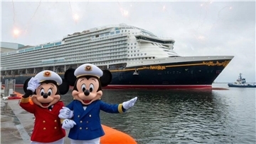 Mickey und Minnie Mouse in Kapitänsuniform posieren vor dem Kreuzfahrtschiff „Disney Adventure“, das im Hafen liegt; im Hintergrund ist ein Schlepper zu sehen.