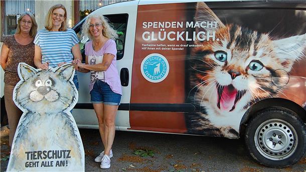 Drei Frauen stehen vor einem Transporter mit der Aufschrift „Spenden macht glücklich“ und einem großen Katzenmotiv. Sie halten ein gemaltes Pappschild mit der Aufschrift „Tierschutz geht alle an!“ in Form einer Katze.