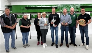 Acht Personen stehen in einer Schießhalle nebeneinander und halten Trophäen, einen Pokal und eine Ehrenscheibe