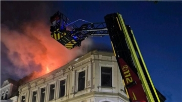 Flammen schlagen aus einem Haus im Hintergrund. Im Vordergrund ist die Drehleiter der feuerwehr zu sehen.