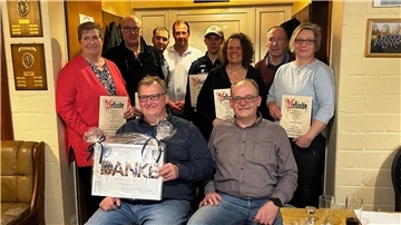 Eine Gruppe von zehn Personen posiert für ein Foto in einem gemütlich eingerichteten Raum mit Holzwänden und Auszeichnungen an der Wand. Mehrere Personen halten Urkunden in den Händen, während ein Mann im Vordergrund eine große, dekorierte Geschenkbox mit der Aufschrift "DANKE" präsentiert. 