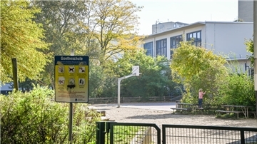 Blick über einen grünen Mattenzaun auf einen Schulhof mit vielen Büschen und einem Basketballkorb. Im Vordergrund steht ein Spielplatz-Schild mit der Aufschrift Goetheschule und sechs Piktogrammen.