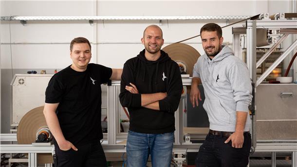Die Gründer des Start-up Papair Fabian Solf (l-r), Christopher Feist und Steven Widdel stehen vor der Maschine, die Luftpolsterfolie aus Papier herstellt.