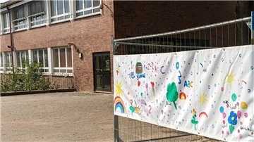 Gebäude mit einem Bauzaun im Vordergrund. An den Gitterstäben des Zauns hängt ein weißes Transparent, das mit vielen bunten Kinderzeichnungen bemalt ist.