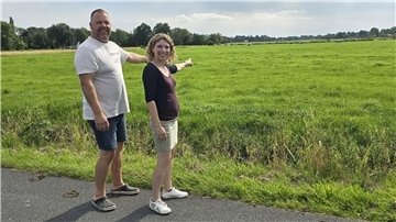 Ein Mann und eine Frau stehen nebeneinander und zeigen vom Wegesrand aus auf eine freie Grünfläche.