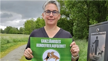 Eine Frau hält einen Zettel in der Hand, auf dem zwei spielende Hunde und der Schriftzug "Ganzjähriges Hundefreilaufgebiet" zu sehen sind. Die Stadt will ihre Freilaufflächen besser kennzeichnen.