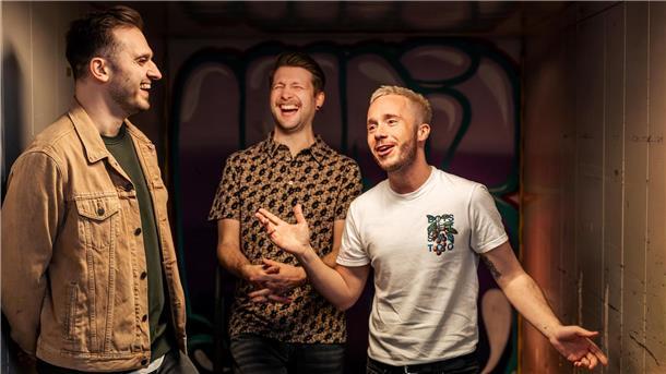 Drei Mitglieder der Band „Von Grambusch“ lachen und unterhalten sich gut gelaunt vor einer farbig besprühten Wand.