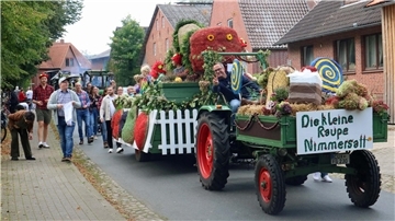 Festlich geschmückter Erntewagen mit dem Motto „Die kleine Raupe Nimmersatt“ fährt durch eine Dorfstraße. Kinder in bunten Kostümen sitzen auf dem Wagen, viele Menschen laufen daneben. Im Hintergrund sind weitere Traktoren und Zuschauer zu sehen.