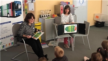 Zwei Frauen sitzen in einem Raum und lesen einer Gruppe von Kindern eine Geschichte vor. Die Frau links hält das Buch "Die kleine Raupe Nimmersatt" in der Hand, während die Frau rechts neben einem Kamishibai (japanisches Erzähltheater) sitzt, in dem eine Illustration der kleinen Raupe Nimmersatt zu sehen ist. Die Kinder sitzen aufmerksam auf dem Boden und hören zu.