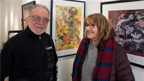 Zwei Personen stehen lächelnd vor gerahmten Kunstwerken in einer Galerie. Links ein älterer Mann mit Brille und schwarzer Jacke, rechts eine Frau mit Brille, brauner Jacke und gestricktem Schal. Im Hintergrund sind farbenfrohe Kunstwerke zu sehen.