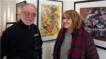 Zwei Personen stehen lächelnd vor gerahmten Kunstwerken in einer Galerie. Links ein älterer Mann mit Brille und schwarzer Jacke, rechts eine Frau mit Brille, brauner Jacke und gestricktem Schal. Im Hintergrund sind farbenfrohe Kunstwerke zu sehen.