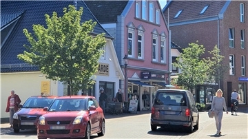Auf einer kleinen Einkaufsstraße fahren drei Autos, Menschen gehen auf den Gehwegen.