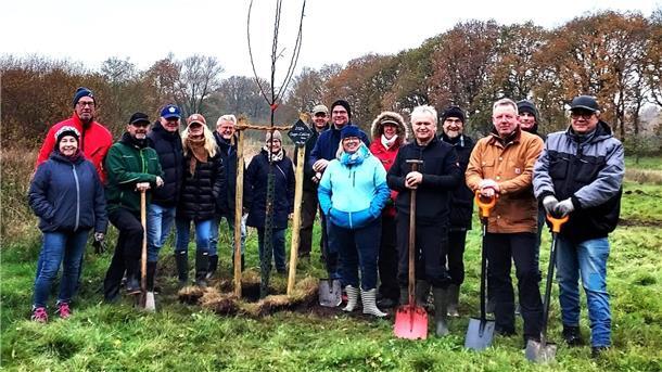 Eine Gruppe von etwa 15 Personen steht auf einer Wiese um einen frisch gepflanzten jungen Baum. Viele der Personen halten Spaten in den Händen und tragen wetterfeste Kleidung. Im Hintergrund sind herbstliche Bäume und ein bewölkter Himmel zu sehen.