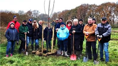 Eine Gruppe von etwa 15 Personen steht auf einer Wiese um einen frisch gepflanzten jungen Baum. Viele der Personen halten Spaten in den Händen und tragen wetterfeste Kleidung. Im Hintergrund sind herbstliche Bäume und ein bewölkter Himmel zu sehen.