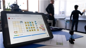 Ein Tablet in einem Klassenzimmer, im Hintergrund ein Schüler und ein Lehrer an der Tafel.