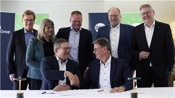 Vertreter von Arla Foods und der DMK Group sitzen an einem Tisch und geben sich nach der Vertragsunterzeichnung lächelnd die Hand, im Hintergrund stehen weitere Führungskräfte vor Arla- und DMK-Logos.