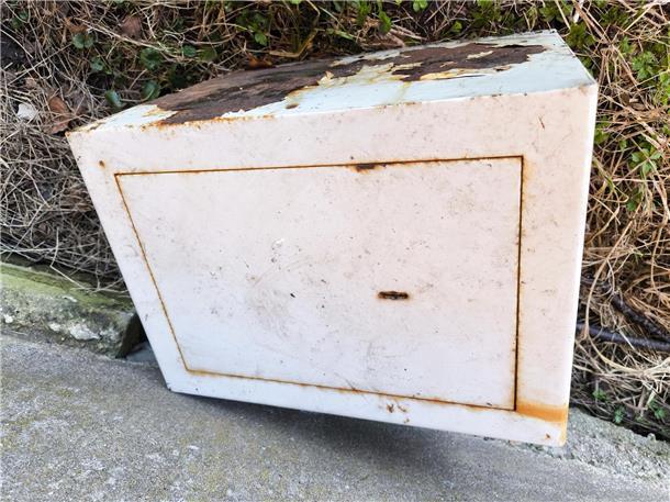 Ein weißer Tresor liegt auf einem asphaltierten Weg. Rostige Stellen sind zu sehen. 