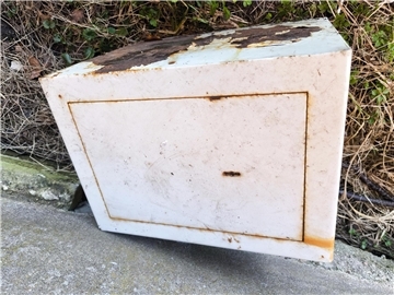 Ein weißer Tresor liegt auf einem asphaltierten Weg. Rostige Stellen sind zu sehen. 