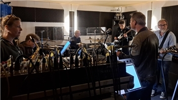 Mehrköpfige Band bei der Probe in einem schallgedämmten Raum mit Schlagzeug, Gitarren, Mischpult und Mikrofonen; konzentrierte Atmosphäre im Tonstudio.