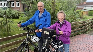 Susanne und Rudolf Nintzel stehen mit Fahrrädern und Fahrradhelmen auf einem gepflasterten Weg, schauen lächelnd in die Kamera. Im Hintergrund ein Garten mit Bäumen und einem Backsteinhaus.