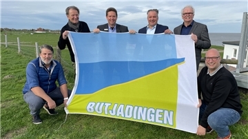 Die Regenbogen-Chefs Marc Voßhall und Rüdiger Voßhall, Bürgermeister Axel Linneweber, Robert Kowitz als Geschäftsführer der Butjadinger Tourismusgesellschaften und Ralph Krümpelmann als deren stellvertretender Aufsichtsratsvorsitzender sowie Regenbogen-Regionalmanager Stephan Jürgens (von links) präsentieren die Flagge, die es für die neuen Camping-Betreiber als Willkommensgeschenk gab.