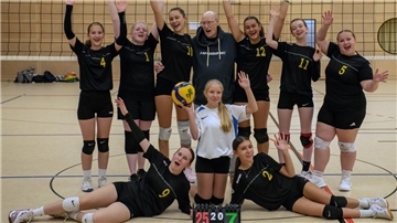 Volleyballerinnen und ihr Trainer stehen vor dem Netz und jubeln in die Kamera.