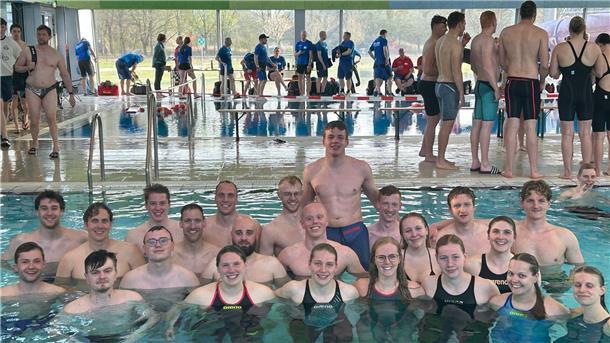 Gruppenfoto von rund 25 jungen Erwachsenen der DLRG Hagen im Bremischen, die in Badekleidung im Schwimmbecken stehen und lächeln; im Hintergrund weitere Wettkampfteilnehmende am Beckenrand einer Schwimmhalle.