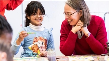 Ein Mädchen mit Brille und Pinsel in der Hand sitzt an einem Tisch und lächelt, während eine Frau in rotem Pullover neben ihr sitzt und ebenfalls lacht. Beide wirken fröhlich und konzentriert beim Malen. Auf dem Tisch liegen bunte Bilder und ein Glas mit Wasserfarbe. 