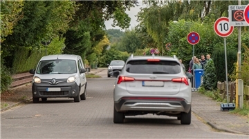 Eine Straße, auf der Autos fahren. Im Hintergrund ist ein Tempo-10-Schild zu sehen. Außerdem ein Tempotafel mit einem traurigen Smiley.