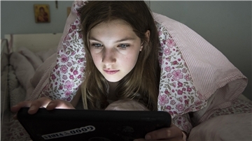 Ein Mädchen liegt auf dem Bett unter der Decke und schaut auf ein Tablet.