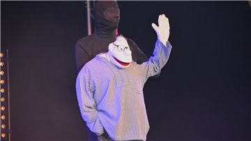 Ein Puppenspieler steht in schwarz gekleidet auf der Bühne und hält eine Stoffpuppe. 