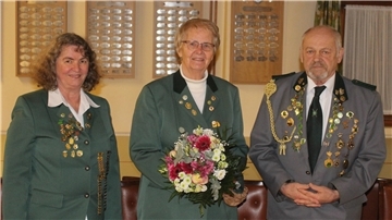 Gruppenfoto mit drei Personen in grüner und grauer Schützenuniform, dekoriert mit Abzeichen. Die Frau in der Mitte hält einen Blumenstrauß, im Hintergrund sind Ehrentafeln an der Wand zu sehen.