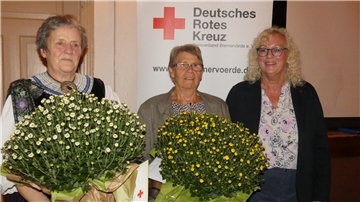 Die DRK-Ortsvereinsvorsitzende Ilse Carstens-Rillox lächelt, während die beiden geehrten Ingrid Schambeck und Margret Köster Blumensträuße in den Händen halten.