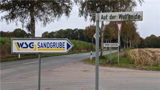 Zwei Wegweiser mit der Aufschrift „WSG Sandgrube“ zeigen auf eine Straße, daneben ein Straßenschild mit dem Namen „Auf der Wulfheide“, im Hintergrund Felder und Bäume.